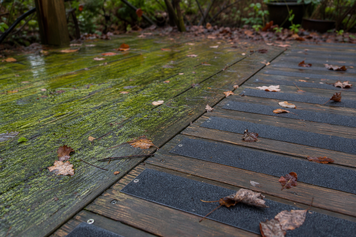 Wet wooden decking with anti-slip strips on the right contrasting with algae-covered, slippery boards on the left, scattered with fallen autumn leaves